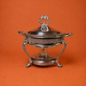 May include: A silver-plated chafing dish featuring a round bowl with a decorative lattice-patterned rim, two side handles, and a matching lid with a floral-shaped handle. The dish is supported by four ornate curved legs and a base.