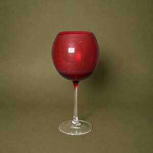 Puede incluir: Una copa de cristal roja con un cuenco redondeado que presenta una serie de finas líneas blancas horizontales grabadas alrededor del exterior. La copa tiene un tallo transparente y delgado y una base circular que aporta estabilidad.
