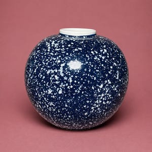 Op de afbeelding: Een bolvormige keramische vaas met een diep marineblauw glazuur en een wit gespikkeld patroon over het oppervlak. De vaas heeft een glad, rond lichaam en een kleine, cirkelvormige opening aan de bovenkant met een strakke witte rand.