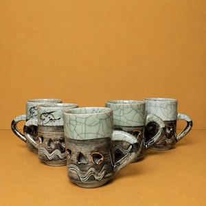 Puede incluir: Un conjunto de cinco tazas de cerámica con un diseño bicolor que presenta un esmalte craquelado verde pálido en la parte superior y una base gris oscuro. La sección inferior muestra un patrón ondulado y recortes en forma de corazón dorados.