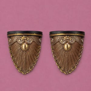 May include: A pair of wall-mounted shelves featuring a semi-circular black top surface and a decorative, fan-shaped base in a metallic bronze finish. The base displays intricate carved details, including a central oval element and scroll accents.
