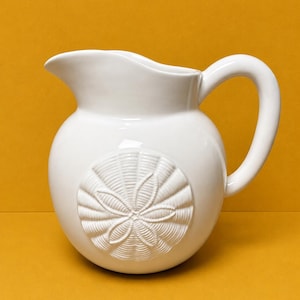 May include: A white ceramic pitcher with a rounded body and a curved handle. The front of the pitcher features a raised, embossed design of a sand dollar with detailed radial lines and a central flower-like pattern on its smooth, glossy surface.