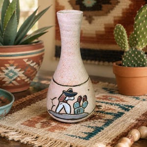 May include: A ceramic vase with a speckled off-white finish and a narrow neck. The body features a hand-painted graphic of a figure wearing a sombrero standing next to a cactus, rendered in blue, brown, and black tones against the light surface.