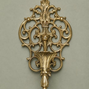 May include: A gold-coloured metal wall sconce featuring an intricate openwork scroll design. The piece includes a central fluted cup-shaped holder supported by a decorative finial at the bottom and an elaborate, symmetrical filigree backplate.