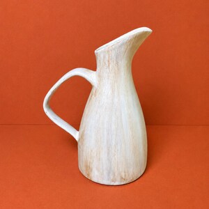 May include: A ceramic pitcher with a tapered body and a curved handle. The surface features a white finish with visible vertical brushstrokes in a light brown or tan hue, creating a textured appearance. The spout is elongated and angled upward.