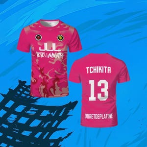 May include: A short-sleeved pink t-shirt featuring a camouflage pattern, the text JUL and TOTO & NINETTA on the front, and the text TCHIKITA, the number 13, and DORETDEPLATINE on the back, displayed from both front and rear perspectives.