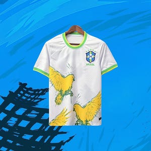 May include: A white short-sleeved t-shirt featuring a yellow bird graphic print, a green and yellow ribbed crew neckline, and the blue and green CBF Brasil logo on the upper left chest. The fabric has a subtle geometric texture throughout.
