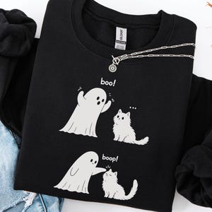 May include: A black long-sleeve sweatshirt featuring a white graphic print of a ghost and a cat. The top panel shows the ghost saying boo! to the cat. The bottom panel shows the ghost touching the cat's head with the text boop! printed below.