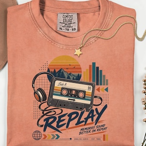 May include: A peach-colored t-shirt featuring a graphic of a cassette tape, headphones, and a sunset over mountains. The text reads "REPLAY", "MEMORIES SOUND BETTER ON REPEAT", and "ANALOG AUDIO EST. 1985". The shirt has a Comfort Colors brand label.