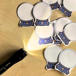 May include: A black flashlight casting a bright beam of light onto a collection of die-cut stickers shaped like crystal balls. Each sticker features a white circular center with a dark blue base decorated with yellow stars and crescent moon icons.