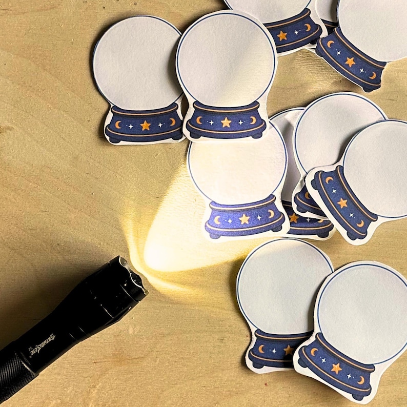 May include: A black flashlight casting a bright beam of light onto a collection of die-cut stickers shaped like crystal balls. Each sticker features a white circular center with a dark blue base decorated with yellow stars and crescent moon icons.