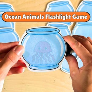 May include: A paper cutout of a light blue fishbowl containing a purple jellyfish with a smiling face. An orange banner at the top displays the text Ocean Animals Flashlight Game in white letters. Multiple identical fishbowl cutouts are visible.
