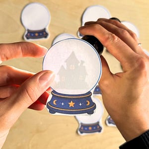 May include: A die-cut paper sticker shaped like a crystal ball with a dark blue base featuring a yellow star and crescent moon design. The clear globe portion displays a light gray silhouette of a house with glowing windows against a white background.