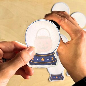 May include: A die-cut sticker shaped like a crystal ball with a blue base featuring a yellow star, crescent moons, and small white stars. Inside the clear globe, a faint orange jack-o'-lantern bucket with a handle is visible as a graphic.