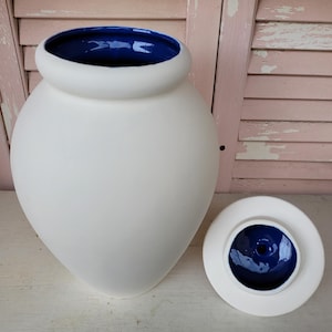 May include: A ceramic jar with a matte white exterior and a glossy deep blue interior. The jar features a rounded body, a wide opening, and a matching removable lid that is white on the top and deep blue on the underside, displayed next to the jar.