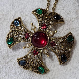 May include: A gold-tone metal cross pendant featuring an intricate filigree design. The center is set with a large round red cabochon, surrounded by smaller square red stones, oval green and blue cabochons, and tiny white faux pearls.