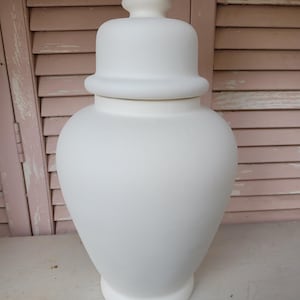 May include: A matte white ceramic ginger jar with a smooth, unglazed finish. The vessel features a rounded body, a tapered base, and a matching removable lid topped with a small, rounded finial. The design is clean, simple, and uniform.