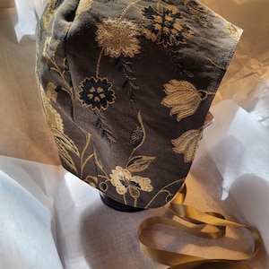 May include: A brown bonnet with gold floral embroidery. The bonnet features a rounded crown and a brim that frames the face. A gold ribbon is attached to the bonnet. The fabric has a textured appearance.