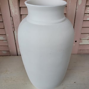 May include: A matte white ceramic vase with a smooth, rounded body and a flared rim. The vessel features a wide, bulbous center that tapers toward a narrow base and a distinct collar detail just below the opening, presenting a clean, uniform surface.