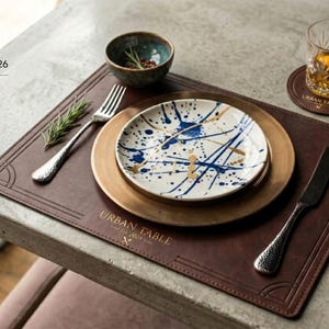 May include: A dark brown rectangular leather placemat with the embossed text URBAN TABLE Est. 2023. It features a round bronze charger plate, a white plate with blue and gold paint splatters, a silver fork, a silver knife, and a matching coaster.