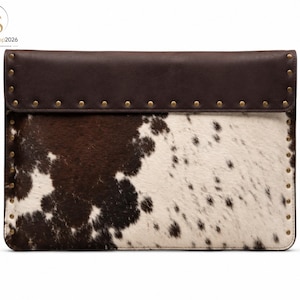 May include: A rectangular clutch bag featuring a brown and white cowhide pattern on the main body and a solid dark brown leather flap. The edges of the flap and the sides of the bag are accented with small, evenly spaced metallic brass studs.