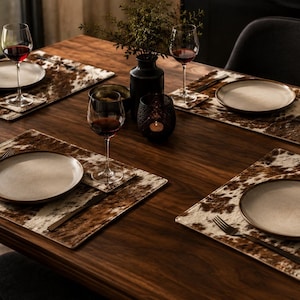 Puede incluir: Una mesa de comedor preparada para cuatro personas con manteles individuales rectangulares con estampado de piel de vaca marrón y blanco. Cada servicio incluye un plato redondo de cerámica color crema, cubiertos de metal y una copa de vino.