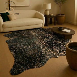 May include: An irregular, cowhide-shaped area rug featuring a mottled pattern of black, dark brown, and cream-coloured spots. The rug has a smooth, flat texture and is positioned on a floor in front of a cream-coloured sofa and an oval coffee table.