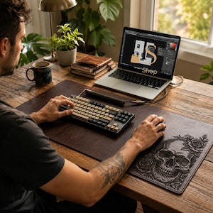 May include: A dark brown leather desk mat featuring an embossed skull and rose design, placed on a wooden desk with a mechanical keyboard, a computer mouse, and a laptop displaying a graphic design interface with the text Shop.