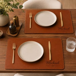 May include: Two rectangular brown leather placemats featuring the name Michael embossed in a script font. Each placemat is set with a white ceramic plate, a gold-toned fork, and a gold-toned knife, arranged on a dark wooden dining table surface.
