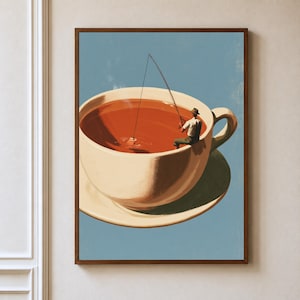 May include: A framed wall art print featuring a large cream-coloured teacup filled with amber liquid against a light blue background. A small figure wearing a hat sits on the rim of the cup, holding a fishing rod with a line extending into the tea.