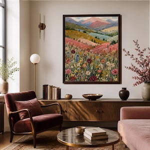 May include: A framed landscape painting featuring a vibrant meadow filled with colorful wildflowers in the foreground, rolling green hills in the mid-ground, and distant pink and blue mountains under a soft sky, displayed on a light wall.