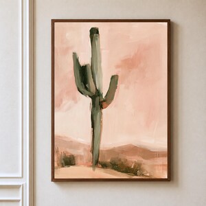 May include: A framed wall art print featuring an abstract painting of a saguaro cactus in shades of sage green against a soft peach and terracotta desert landscape background with visible brushstrokes and a dark brown wooden frame border.