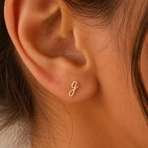 Pode incluir: Um brinco de ouro em forma de letra minúscula g em fonte cursiva. O brinco está posicionado no lóbulo da orelha, apresentando um acabamento metálico polido e um design simples e limpo que assenta diretamente sobre a pele.