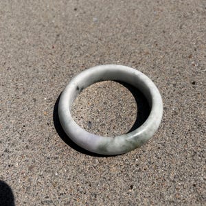 Puede incluir: Un brazalete circular liso hecho de piedra pulida. La pieza presenta una apariencia veteada con una mezcla de tonos blanco translúcido, lavanda suave y verde apagado, mostrando variaciones de color naturales en toda la superficie.