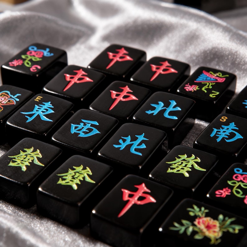 May include: A collection of black Mahjong tiles featuring engraved characters and floral designs in vibrant colors including red, blue, green, and pink. Each rectangular tile has a glossy finish with distinct Chinese characters and symbols.