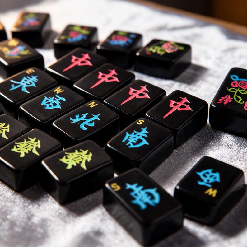 May include: A collection of black rectangular Mahjong tiles featuring engraved characters and symbols in vibrant colors including red, blue, yellow, and green. Each tile displays distinct Chinese calligraphy and directional letters like E, W, N, and S.