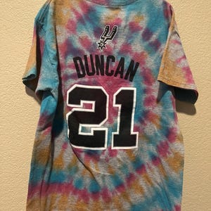 May include: A short-sleeve t-shirt featuring a multi-colored tie-dye pattern in shades of blue, pink, and orange. The back displays the San Antonio Spurs logo, the name DUNCAN, and the number 21 printed in black with a white outline.