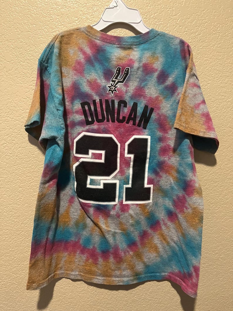 May include: A short-sleeve t-shirt featuring a multi-colored tie-dye pattern in shades of blue, pink, and orange. The back displays the San Antonio Spurs logo, the name DUNCAN, and the number 21 printed in black with a white outline.