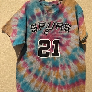 May include: A short-sleeve t-shirt with a tie-dye pattern in shades of blue, pink, and tan. The front features the word SPURS in black and white lettering with a spur logo, and the number 21 in large black and white digits centered below.