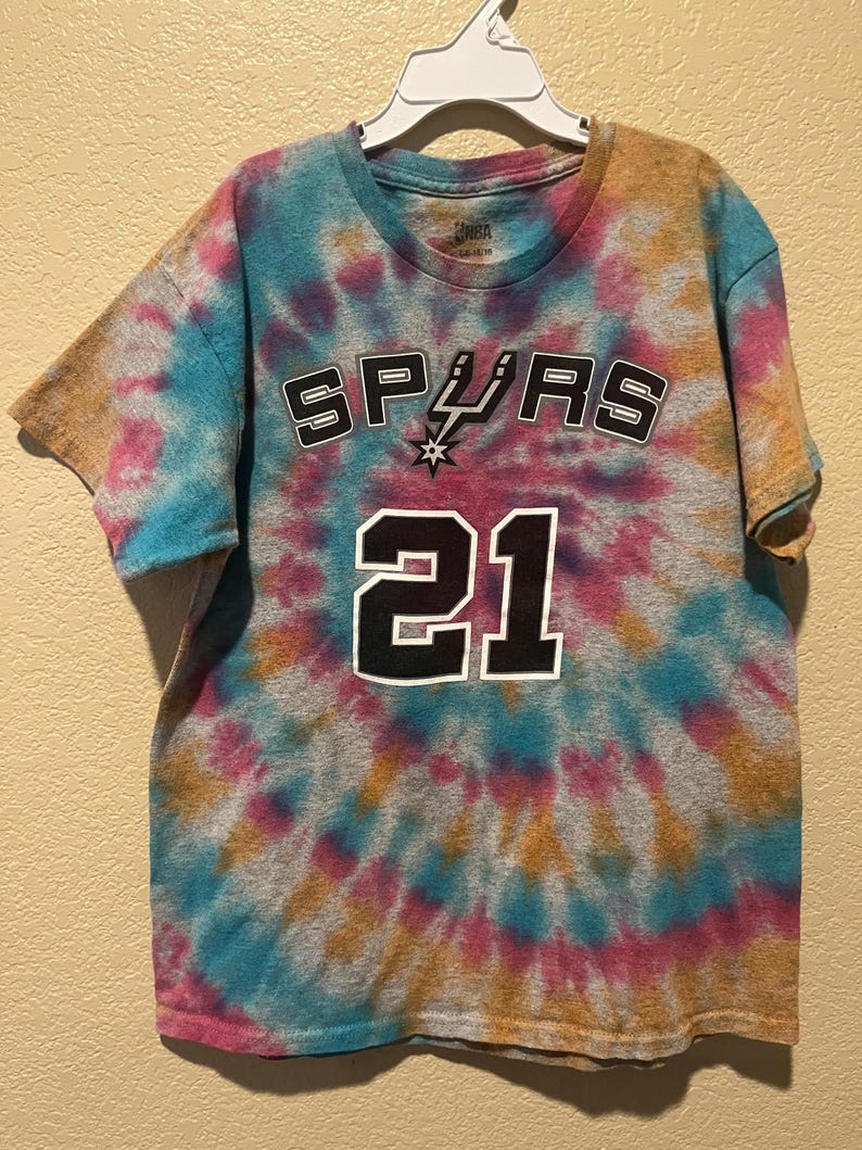 May include: A short-sleeve t-shirt with a tie-dye pattern in shades of blue, pink, and tan. The front features the word SPURS in black and white lettering with a spur logo, and the number 21 in large black and white digits centered below.