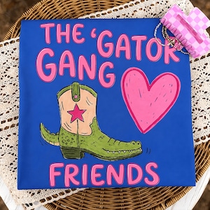 Op de afbeelding: Een blauw t-shirt met de roze tekst THE GATOR GANG FRIENDS, een groene cowboylaars met een krokodillenstaart en een roze ster, en een groot roze hart. Een roze en wit geblokte haarklem met een kralenketting ligt in de rechterbovenhoek.