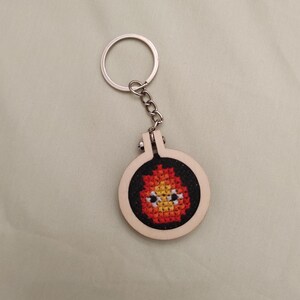 Può includere: Un portachiavi a forma di telaio da ricamo circolare in legno con un disegno a punto croce di un personaggio a fiamma stilizzato arancione, giallo e rosso con due occhi bianchi e pupille nere su sfondo in tessuto nero con anello in metallo.