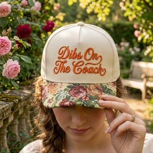 May include: A cream-colored baseball cap featuring the embroidered orange text "Dibs On The Coach" on the front panel. The brim of the cap is decorated with a colorful floral pattern consisting of red, pink, and yellow roses on a light background.