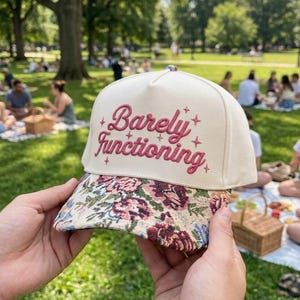 May include: A cream-colored baseball cap featuring the embroidered text "Barely Functioning" in dark pink, accented by small star symbols. The brim of the cap displays a colorful floral tapestry pattern with shades of pink, green, and blue.