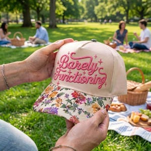 May include: A beige baseball cap featuring the embroidered pink text Barely Functioning with small star accents. The brim of the cap displays a colorful floral pattern with red, yellow, purple, and green flowers against a light background.