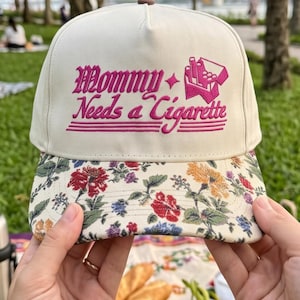 May include: A cream-colored baseball cap featuring an embroidered pink graphic that reads "Mommy Needs a Cigarette" with a small cigarette pack icon. The brim is decorated with a colorful floral pattern in shades of red, yellow, blue, and green.