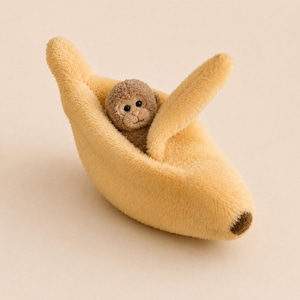 May include: A plush toy featuring a small, brown, fuzzy monkey nestled inside a soft, yellow, banana-shaped peel. The monkey has dark eyes and a light pink nose, while the banana peel has a textured, fuzzy surface with a brown tip at the end.