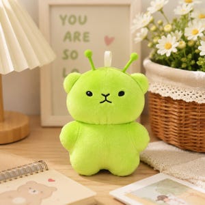 May include: A bright lime green plush toy shaped like a bear with two antennae on its head. The plush has small black embroidered eyes and a simple black stitched mouth. It features a soft, fuzzy texture and a small white fabric loop on top.
