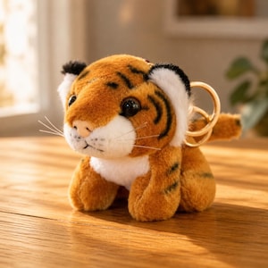 May include: A plush tiger keychain featuring orange fur with black stripes, a white muzzle, and white inner ears. The stuffed animal has large black eyes, thin white whiskers, and a gold-toned metal ring attached to the back for hanging.
