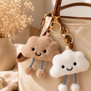 May include: Two plush cloud keychains attached to a gold-toned metal ring. One cloud is light brown and the other is white. Both feature embroidered black eyes and a smiling mouth, with two dangling legs made of black and white striped cord.